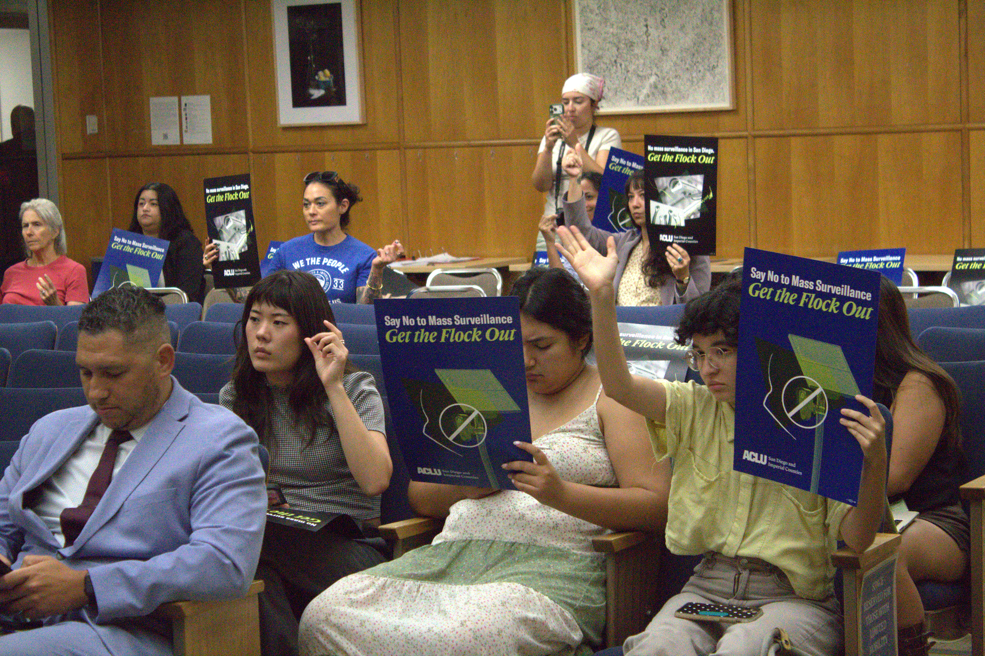 People sitting in chairs at a San Diego City Council meeting hold up signs that say “Say No to Mass Surveillance, Get the Flock Out.” 