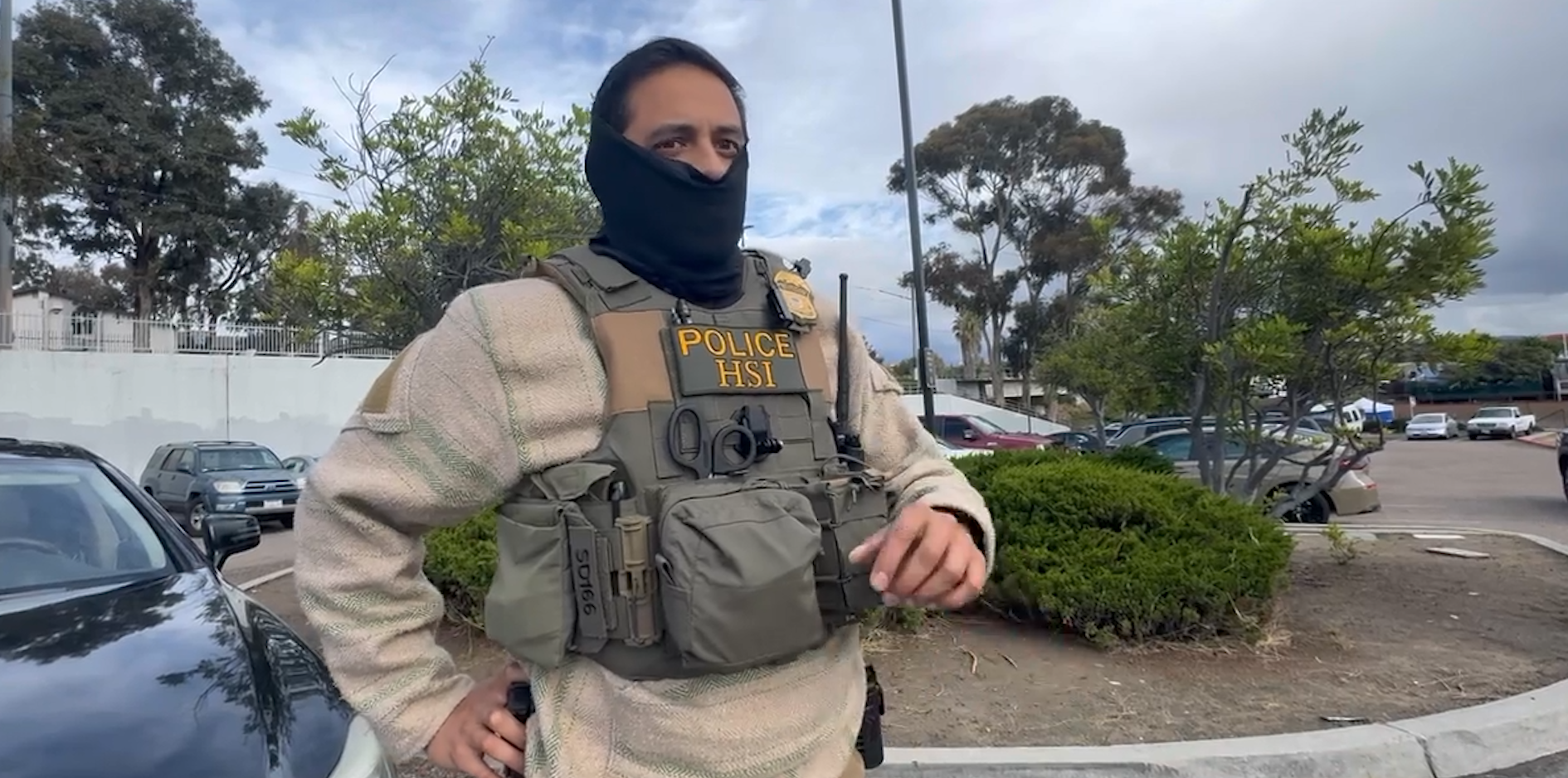 A man with a black face covering, a bulletproof vest that says Police HSI with badge number 50166 stands next to a black car in a parking lot