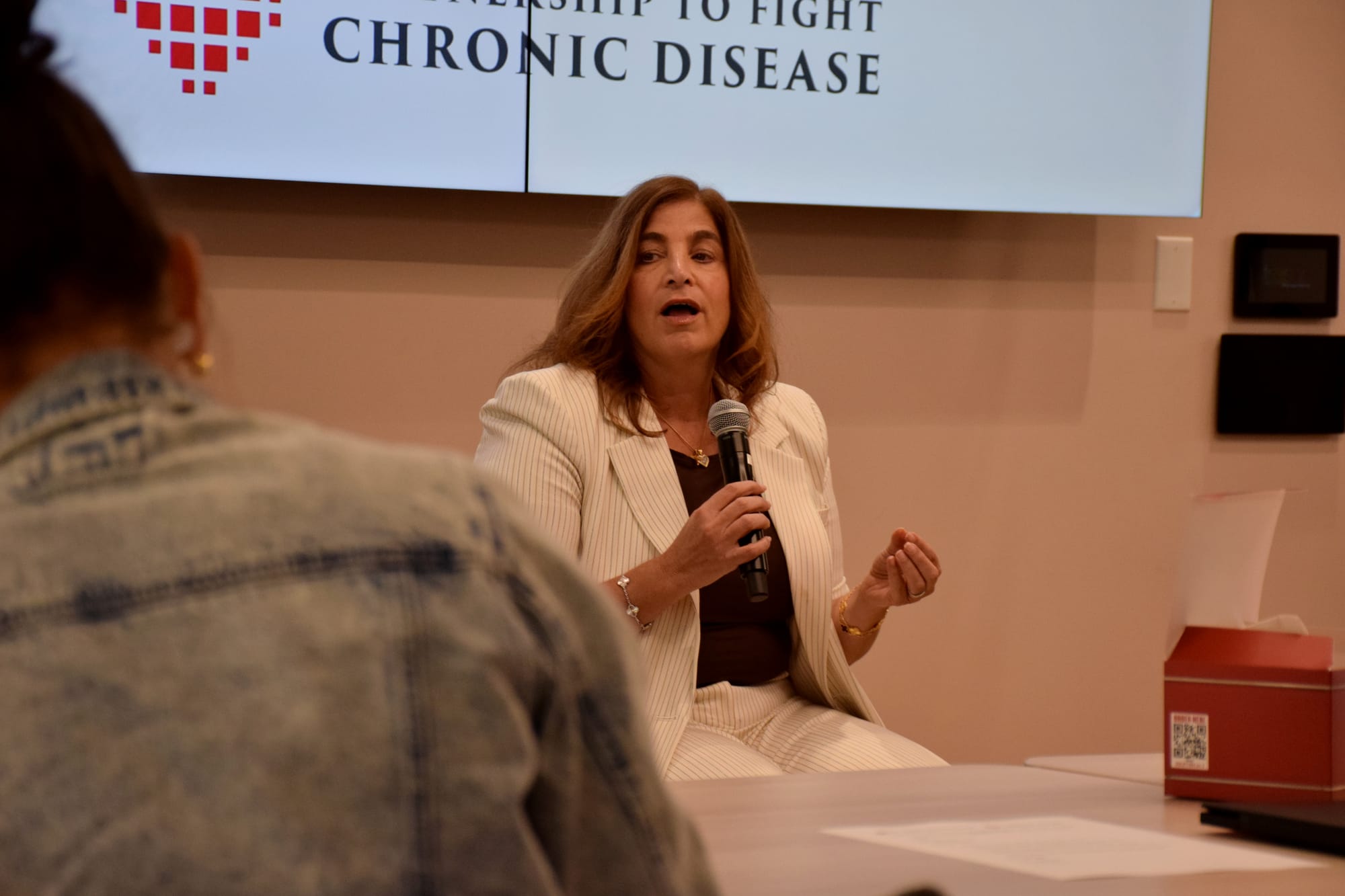 A woman in a white pantsuit speaks into a microphone while sitting at the front of a conference room