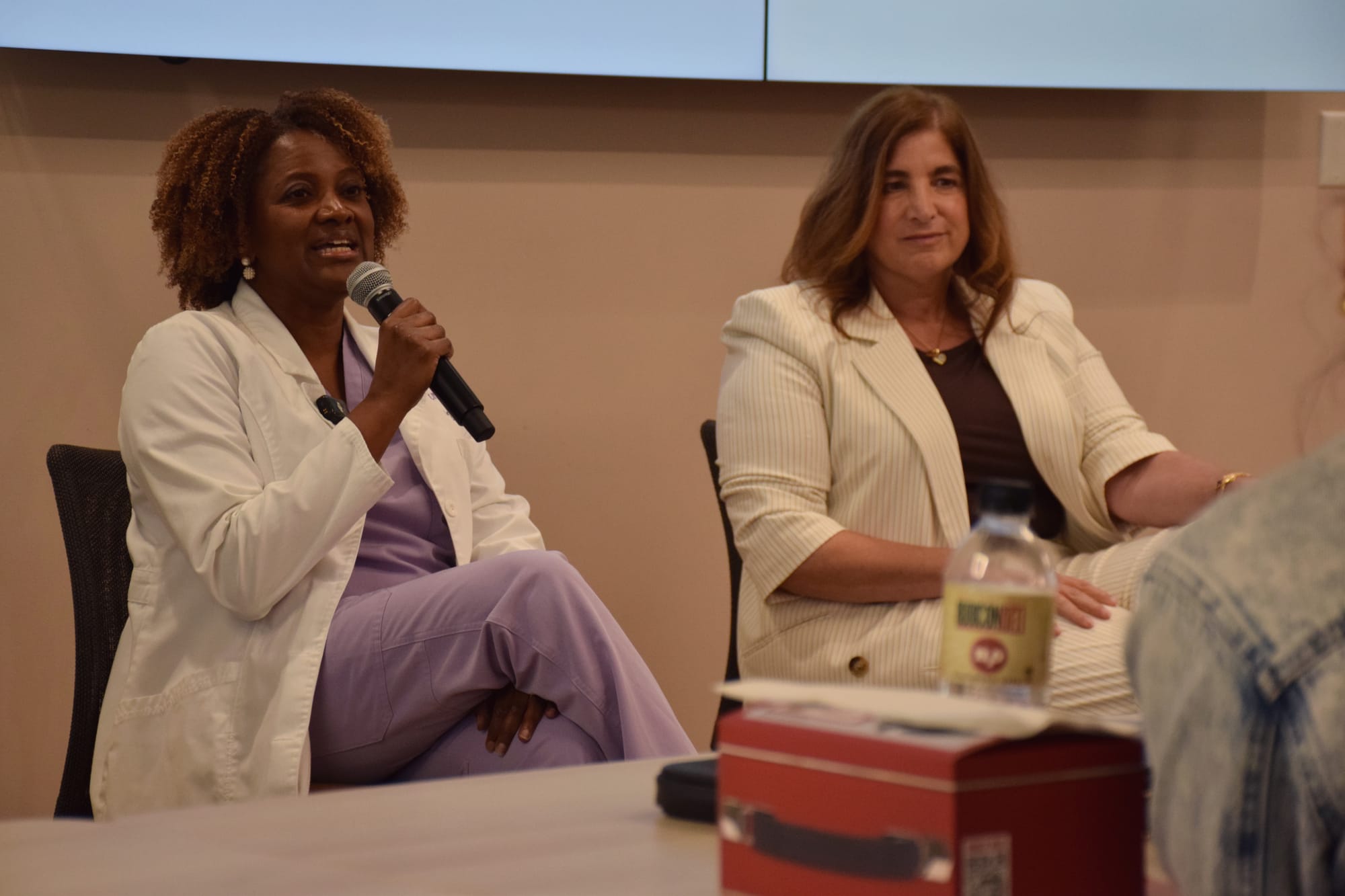 A woman in lilac scrubs and a white lab coat speaks into a microphone sitting next to another woman in a white pantsuit