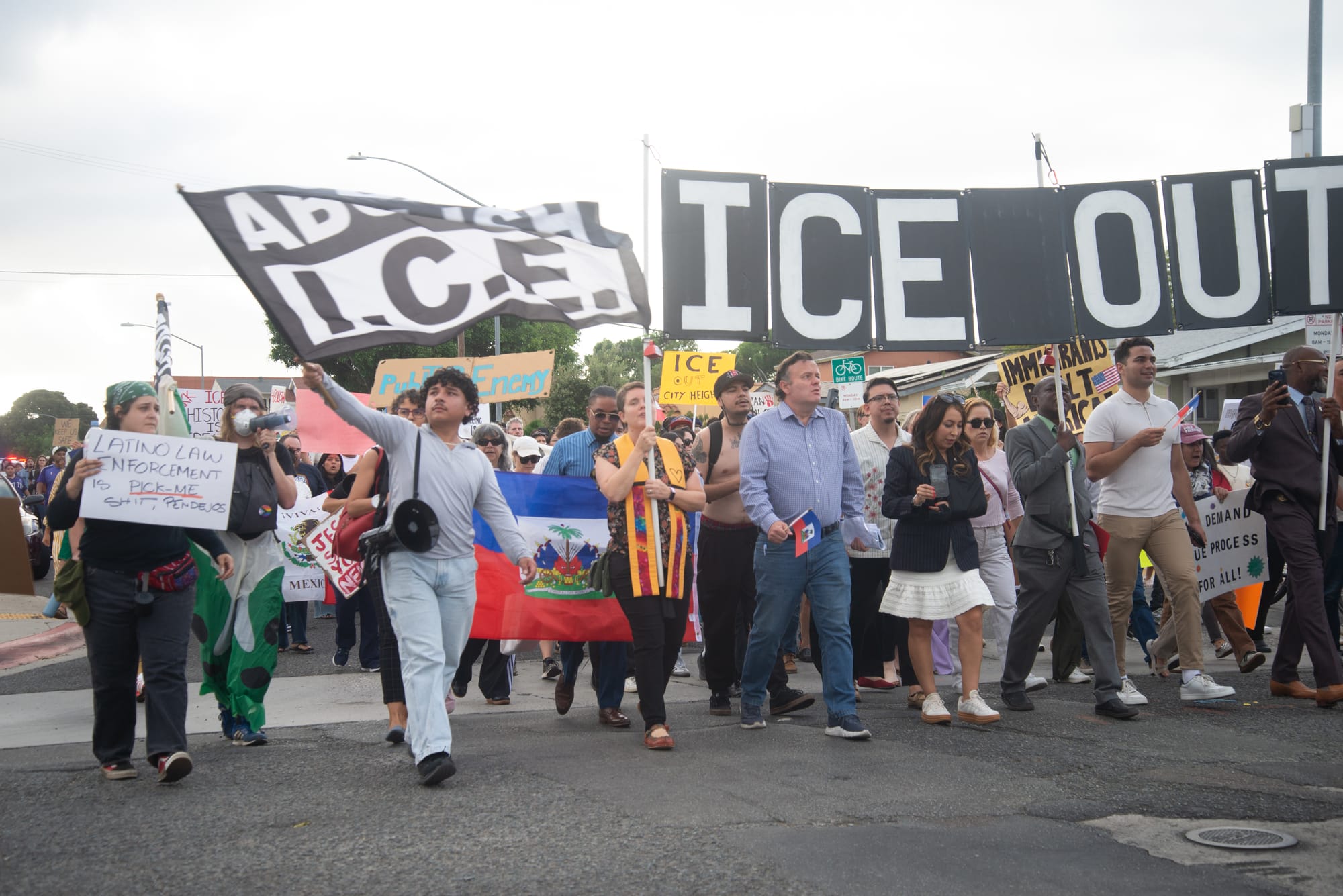 A man wearing gray holds a black flag that says Abolish I.C.E. standing in front of a large group of people