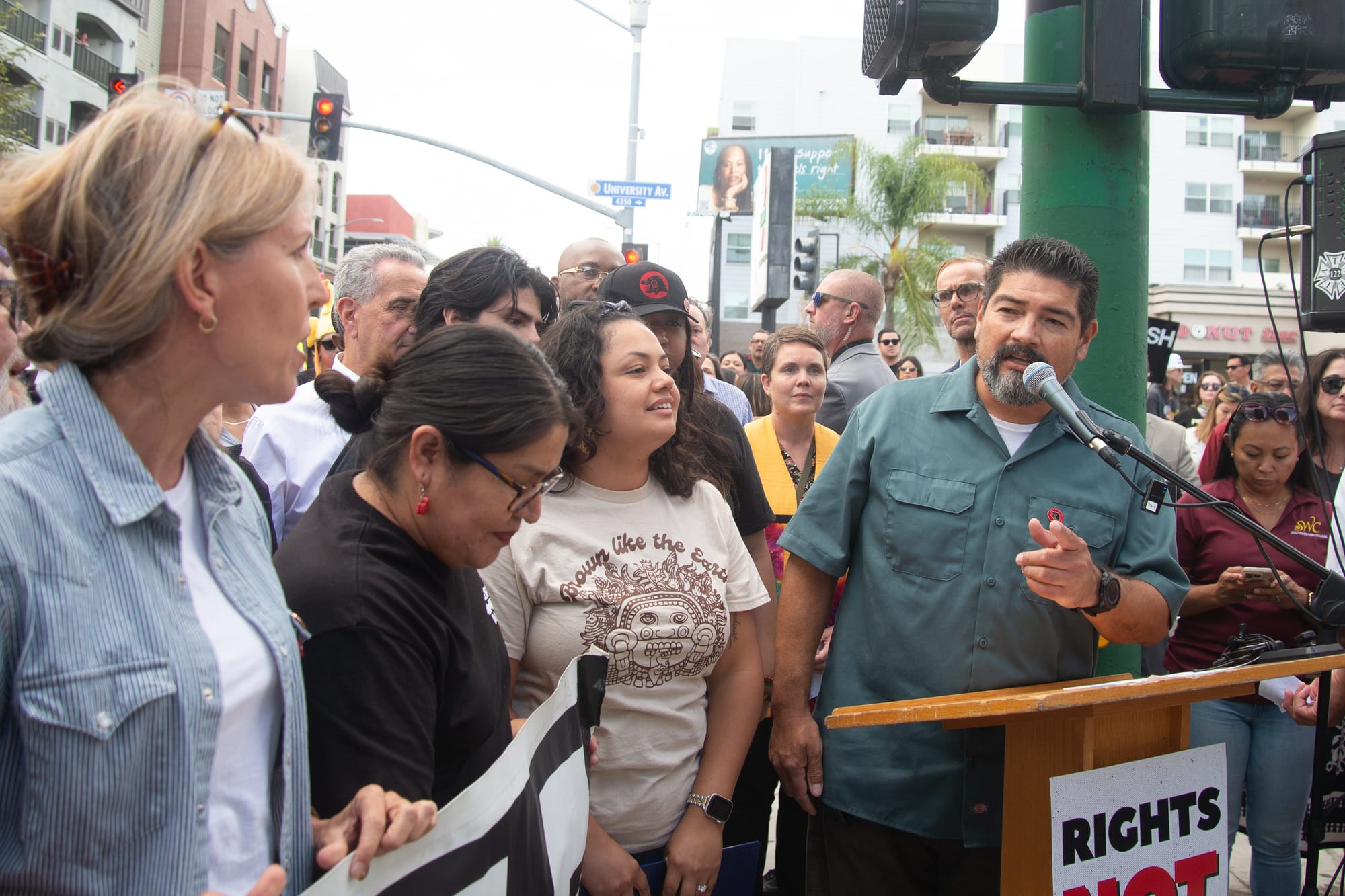 A man in a blue button down shirt speaks at a podium surrounded by people including a woman with a beige shirt that says brown like the earth