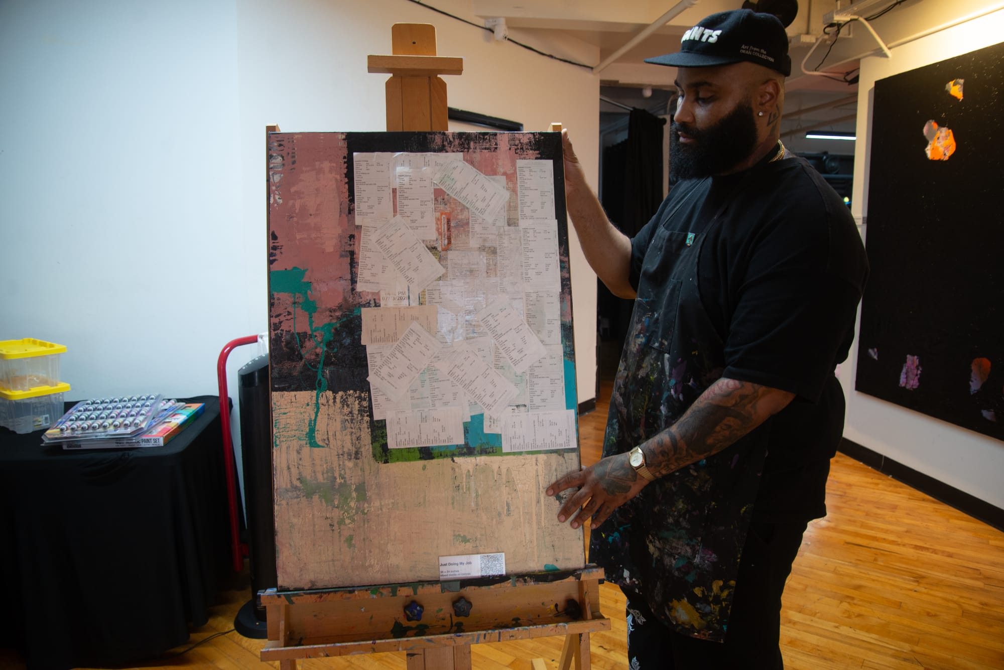 A man in a black hat and black shirt stands next to a painting on an easel that has parking tickets on it