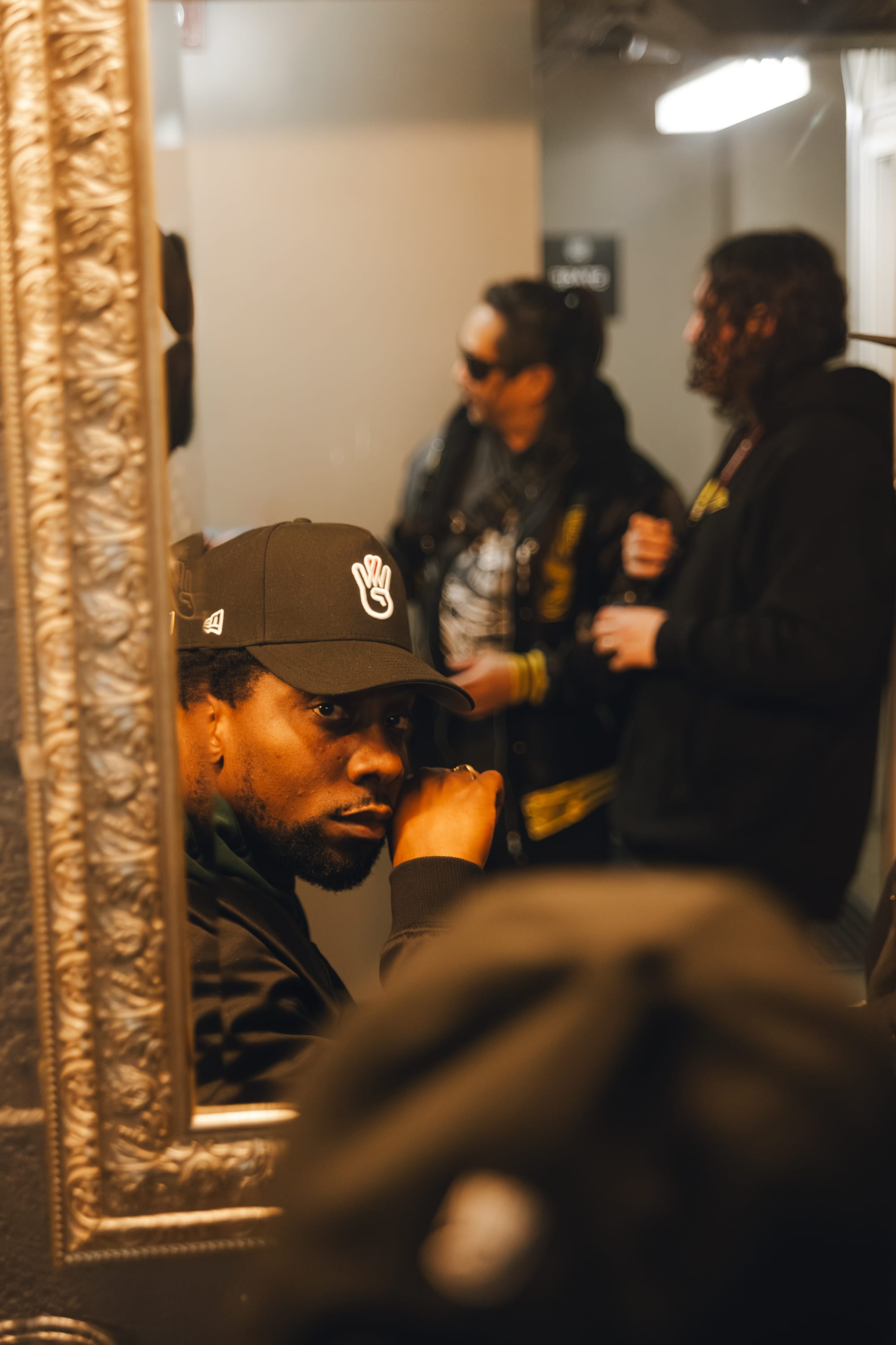 A man in a black hat and jacket looks into a gold framed mirror