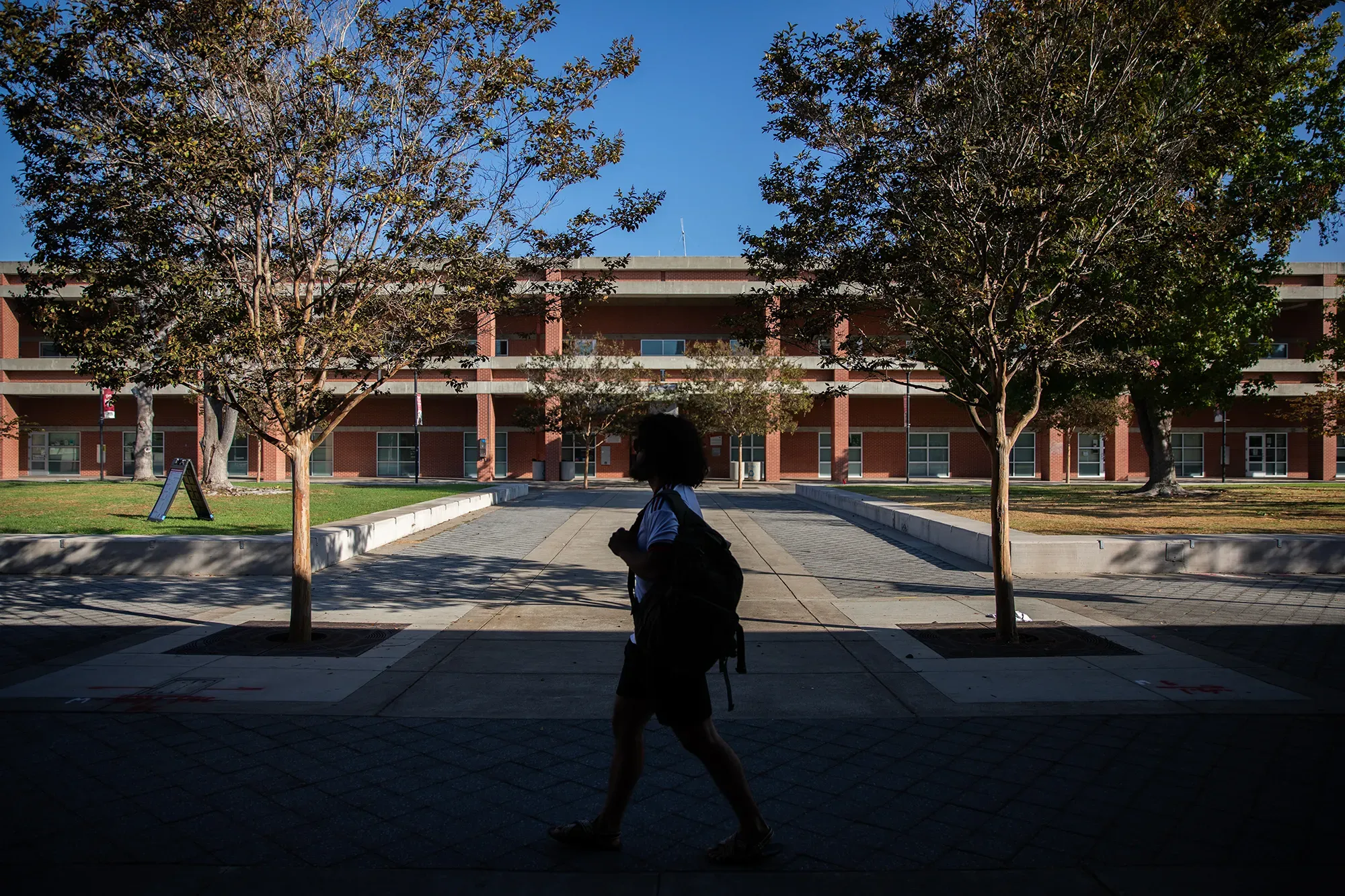 A silhouetted person carrying a backpack walks in front of a brick building with trees