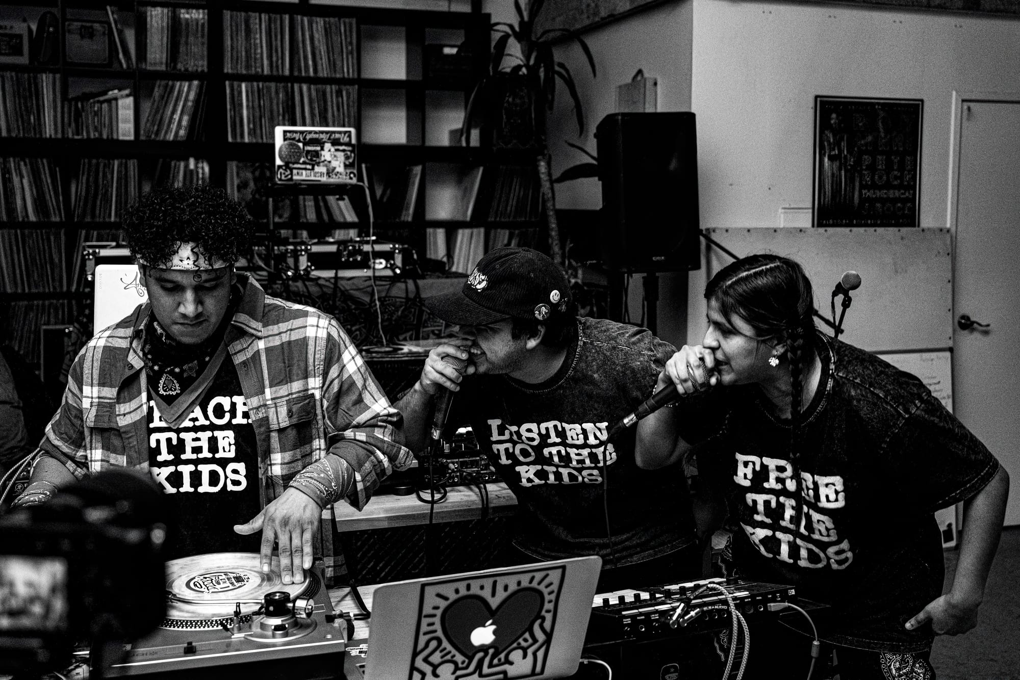 A black and white photo of a DJ and two MCs where both the MCs are watching the DJS work as they hold the mics to their faces