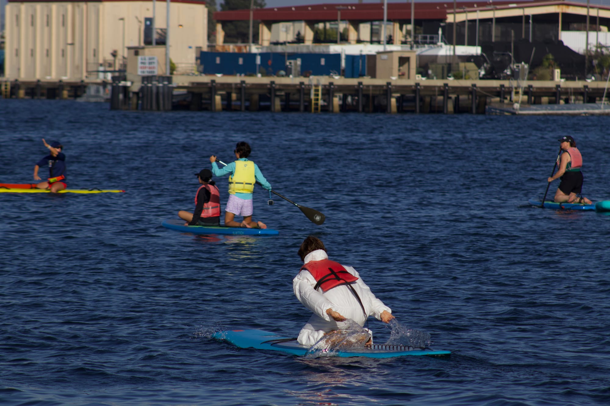 A person in a white suit facing away from the camera swings their arms to paddle a board forward in the water