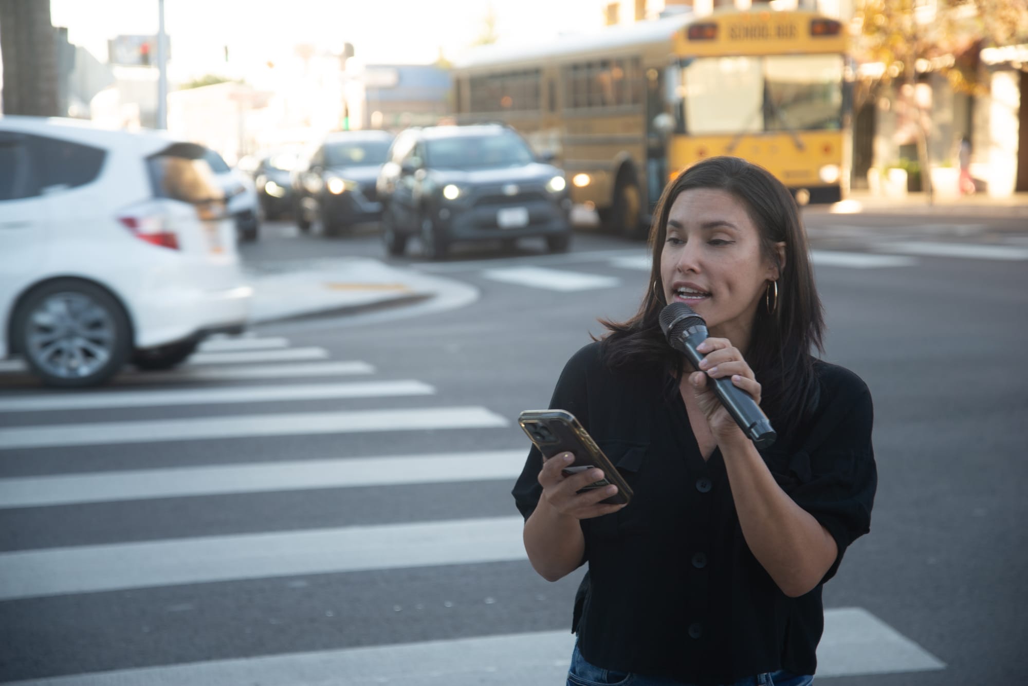 A woman in a black shirt holds a microphone while standing on a street corner