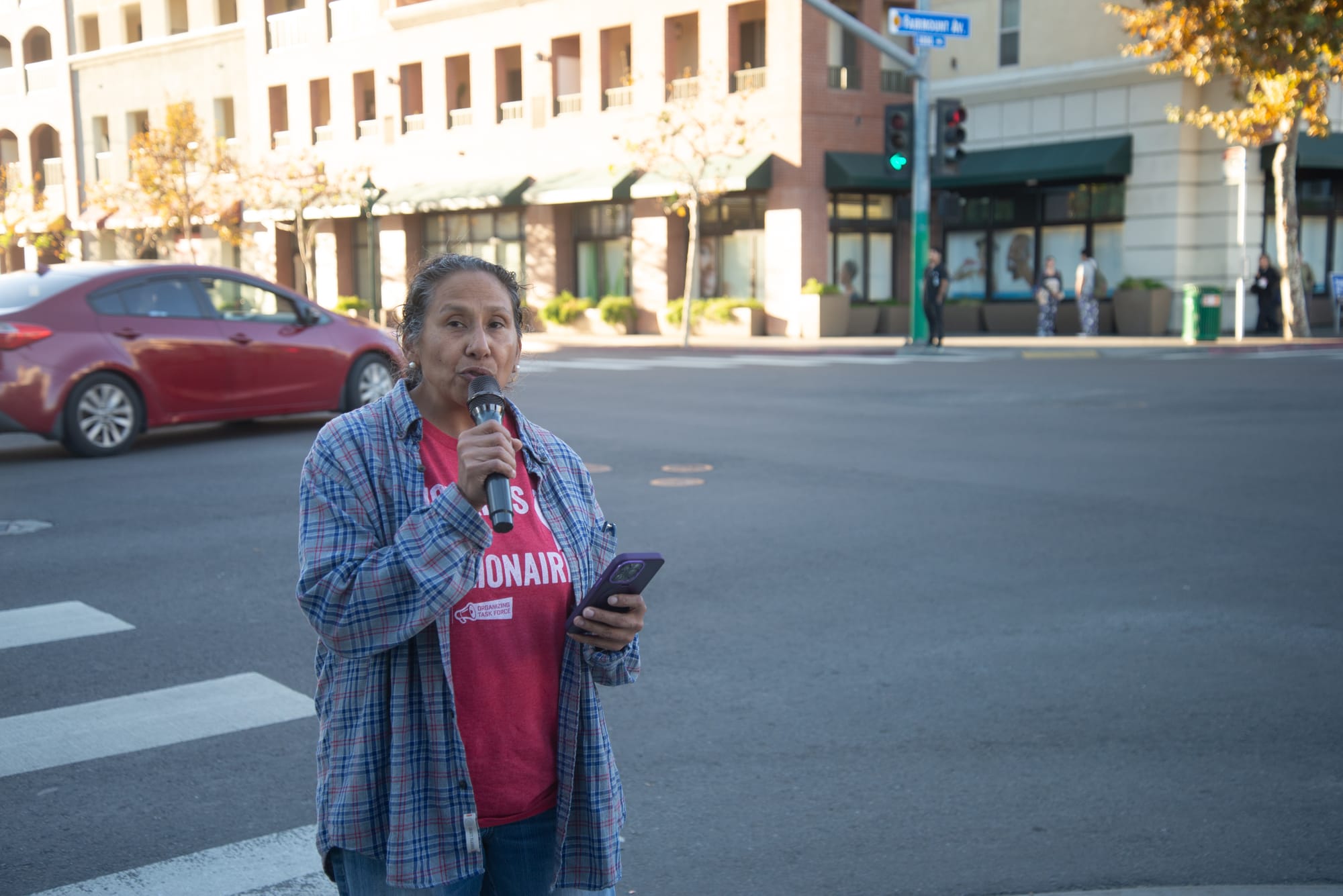 A woman in a red shirt and plaid button down holds a microphone while standing on a street corner