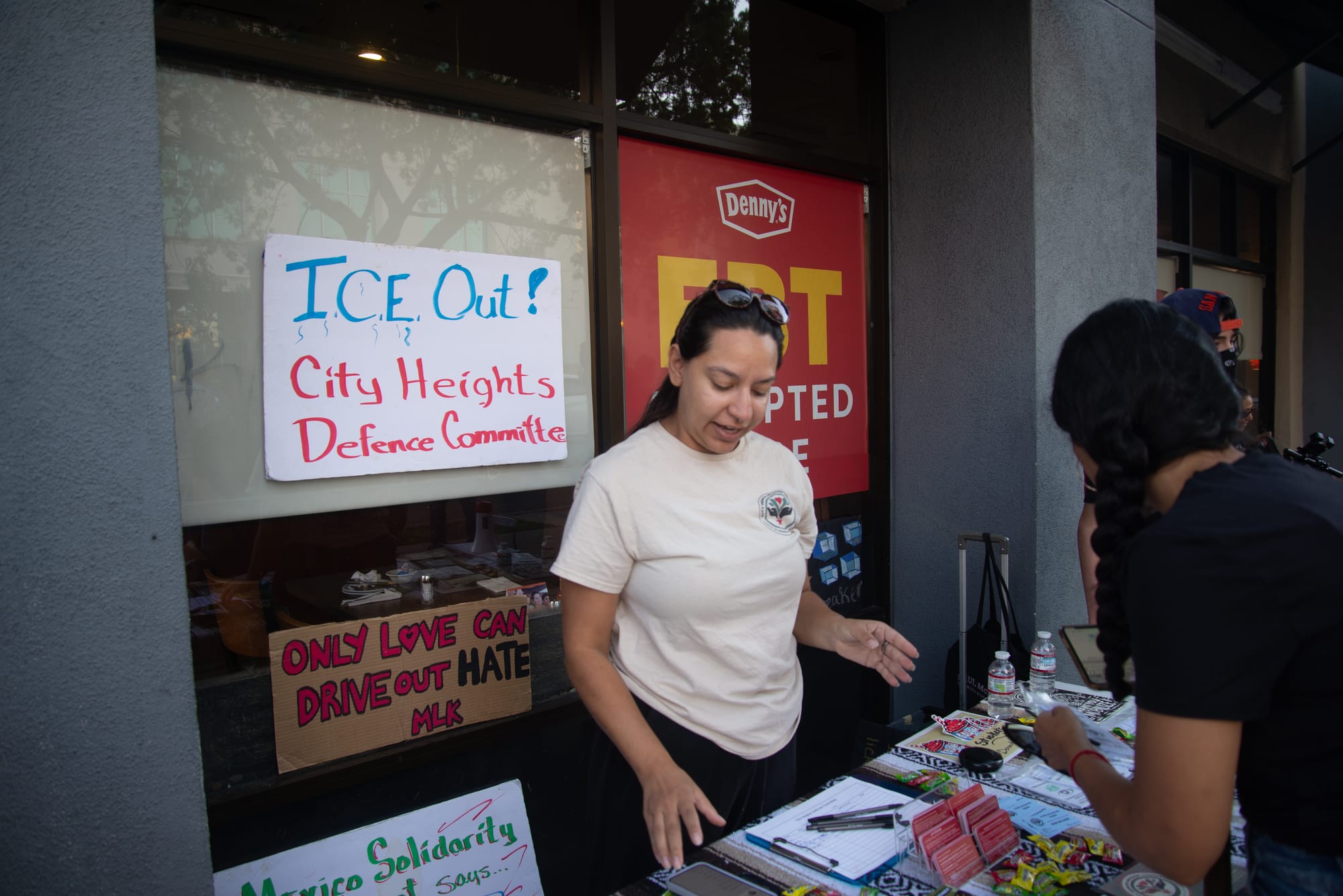A person stands at a table with materials on it and a sign behind that says ICE Out! City Heights Defense Committee