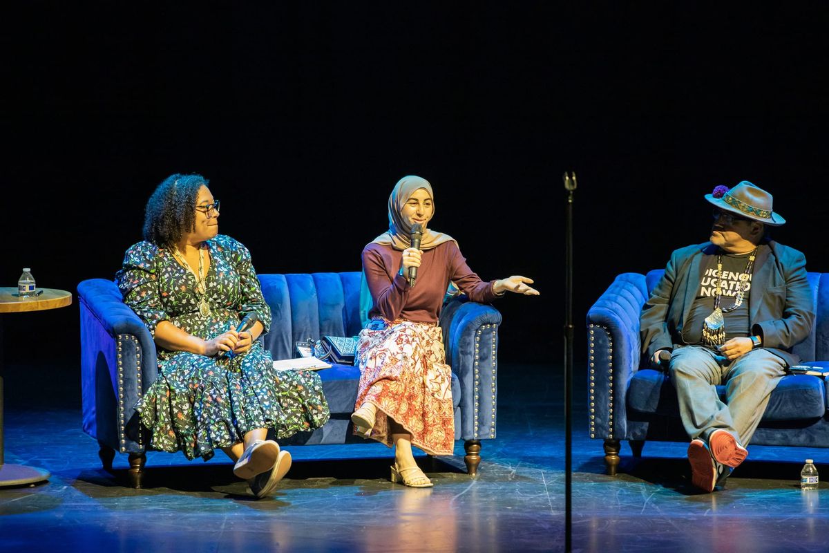 two women sit on a couch, one wearing a dress and the other wearing a skirt and hijab. A man in a hat sits on a couch next to them.