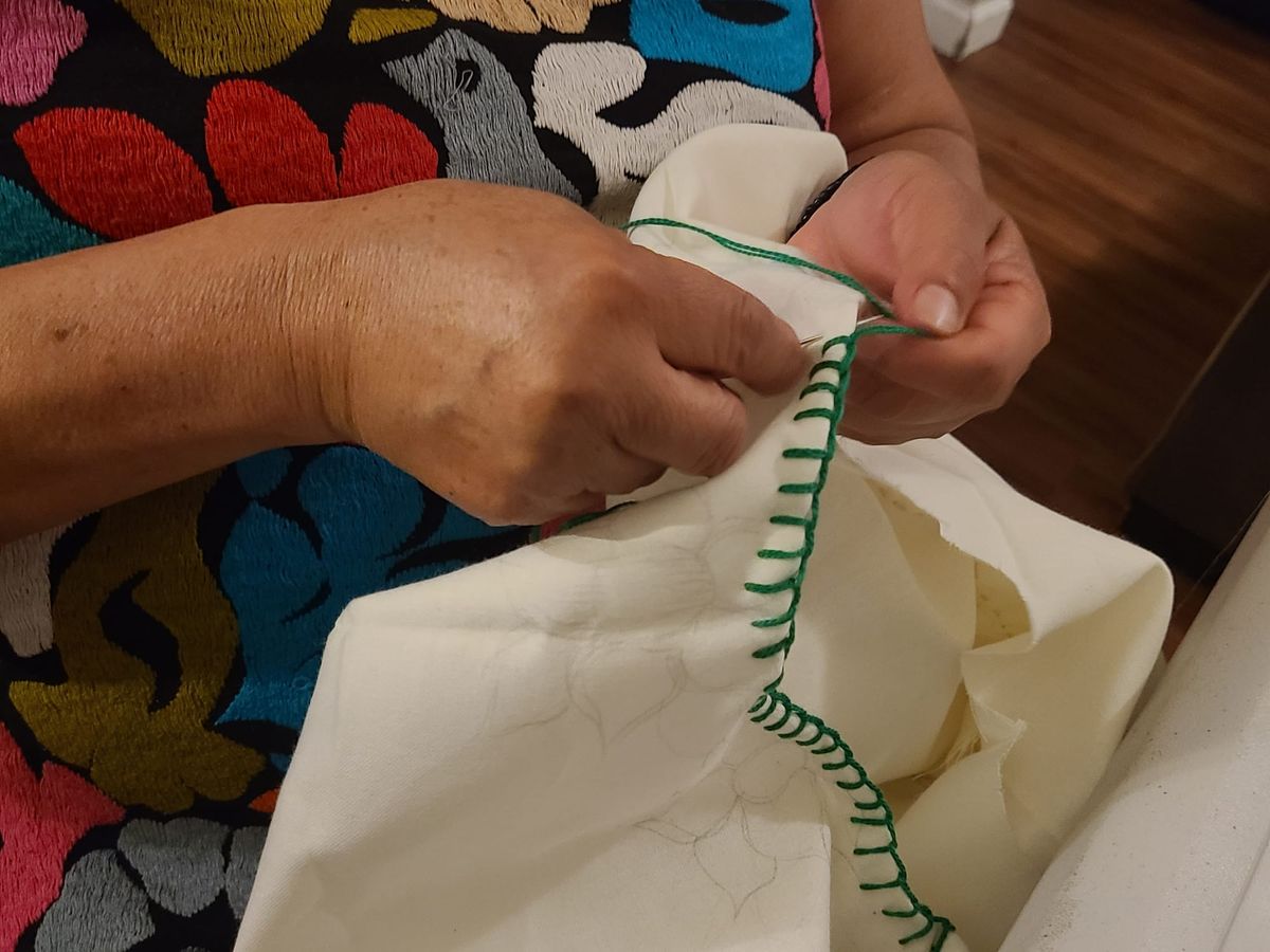 close up of a woman's hands sewing green thread on the edge of a white cloth