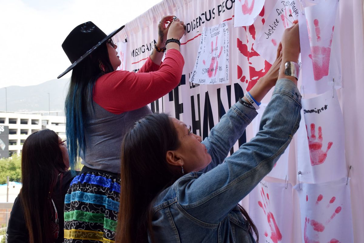 Three women pin cloths with red handprints on them to a large sign at an outdoor event