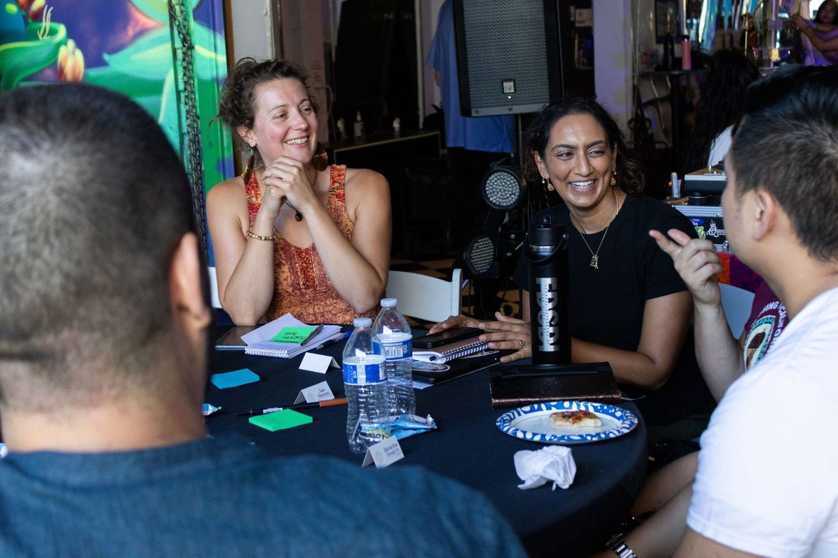 Two women smile at a table facing two people whose backs are to the camera. The table has water bottles and post-it notes on it.