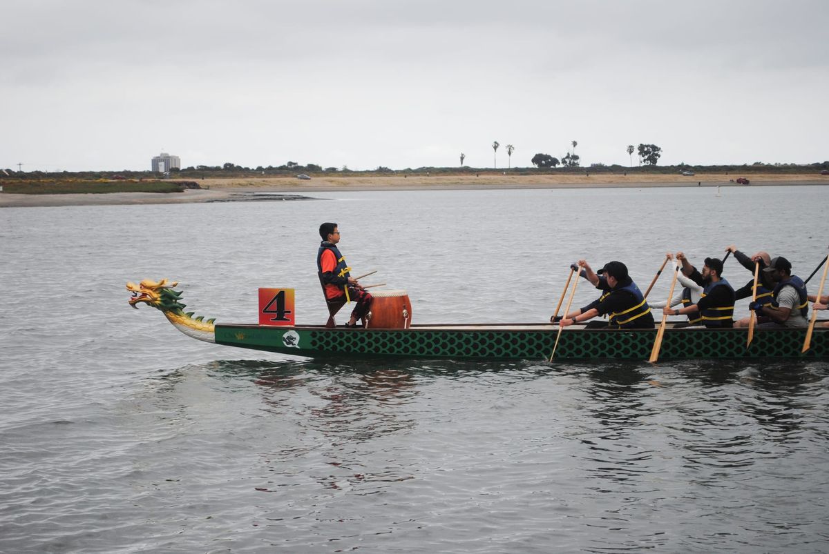 A long slender dragon boat with black and green painted scales moves to the left carrying a drummer on a chair and several paddlers
