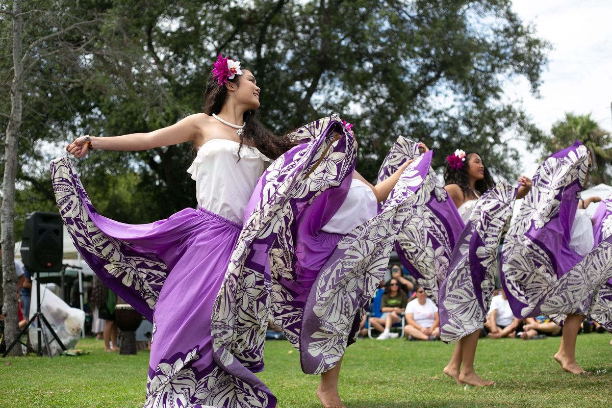 Four women wearing purple, patterned skirts, white shirts and flowers in their hair dance in front of a tree on the grass at an event with people sitting in the background