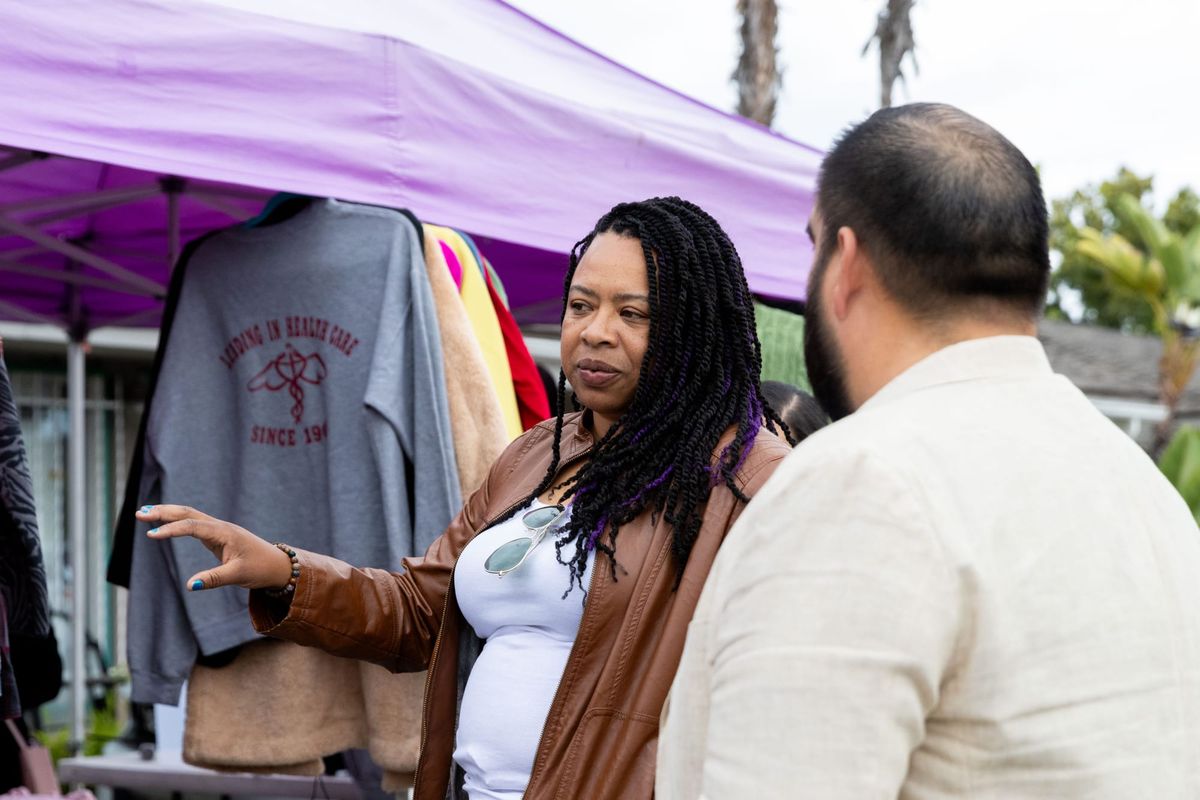 A woman speaks to a man outside in front of clothing hung up under a purple tent