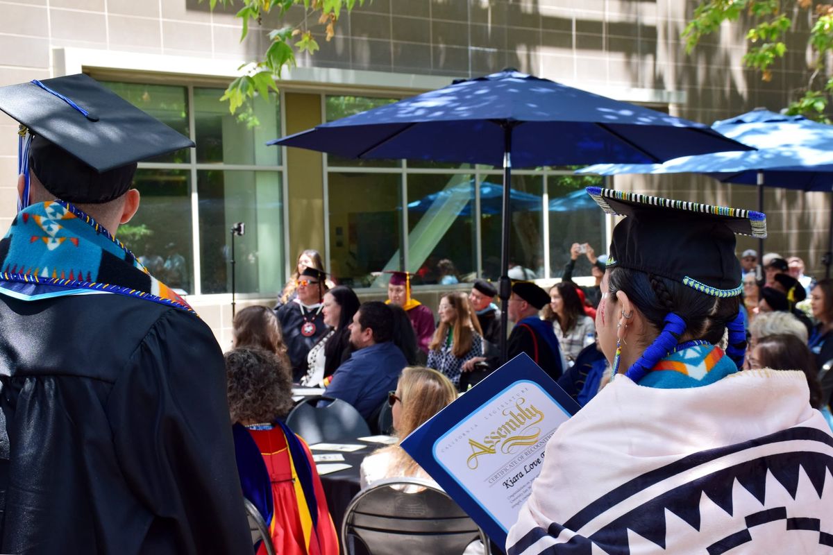 The backs of two graduates in front of a crowd of people at an outdoor event