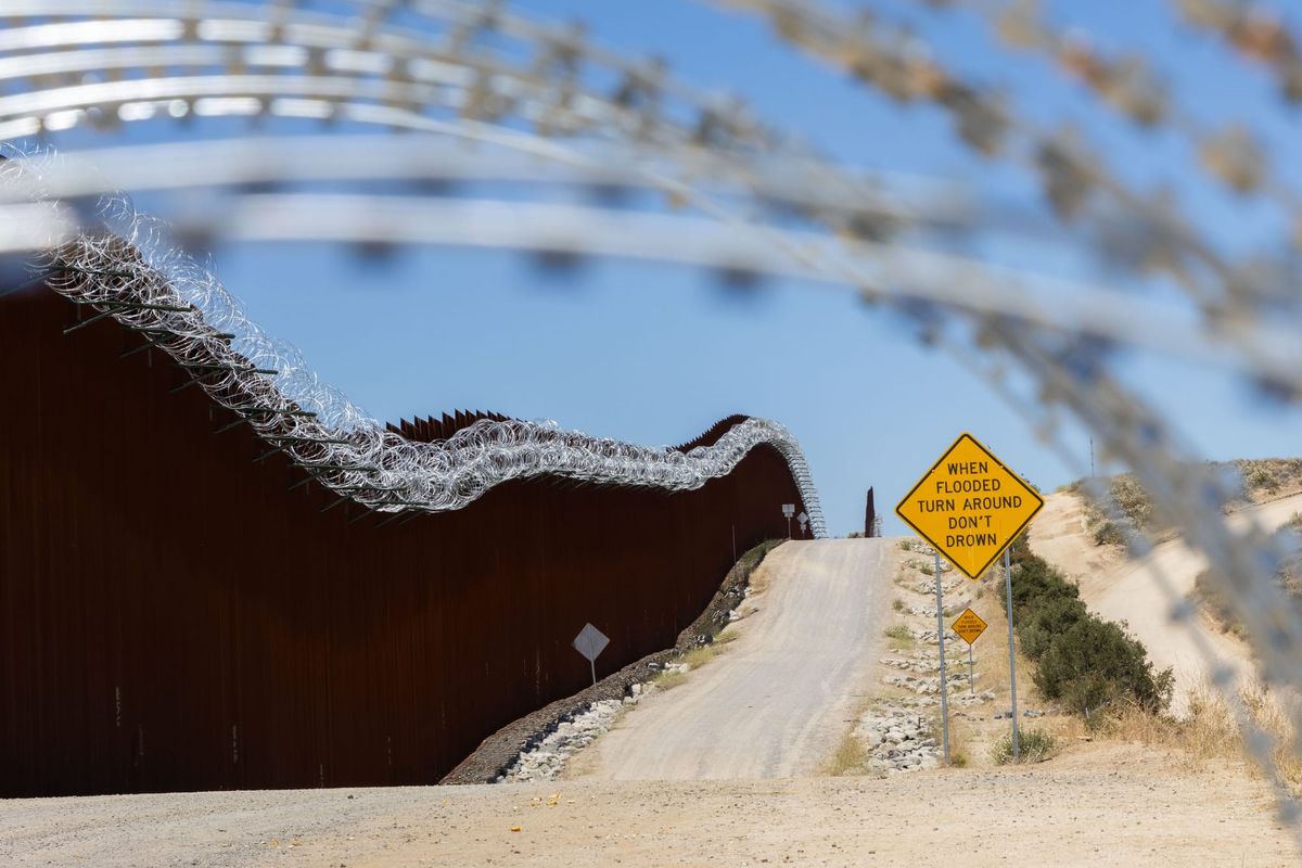 A dirt road runs along a border wall with a sign that says "When flooded turn around don't drown"