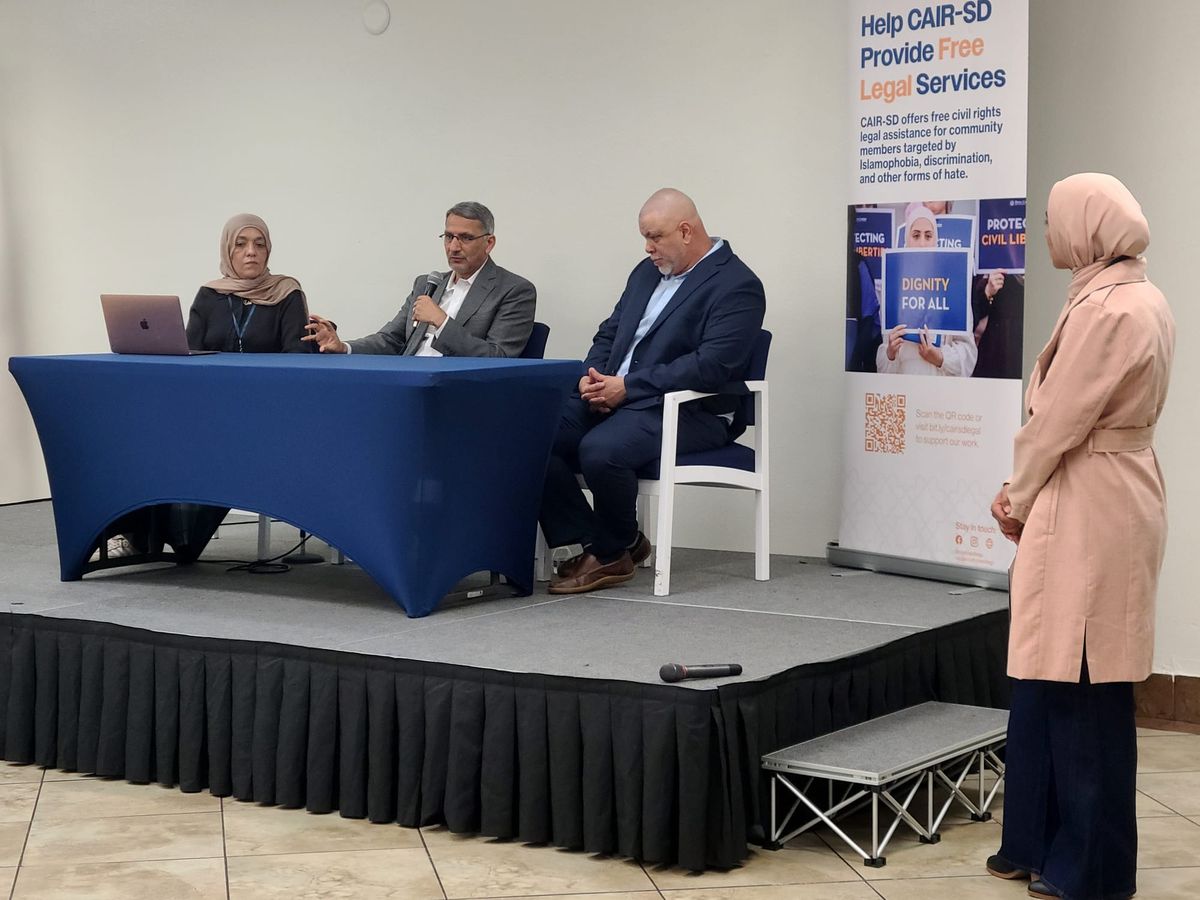 Three people sit on a panel at a conference with one speaking into a microphone 