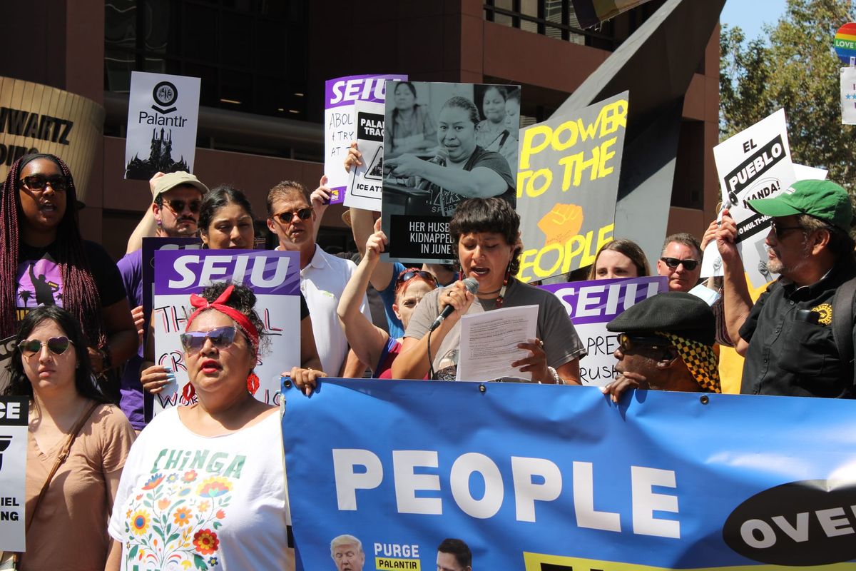 A person speaks into a microphone at a rally with other protesters nearby holding signs reading "Palantir," "Her husband was kidnapped in June", "Power to the People"