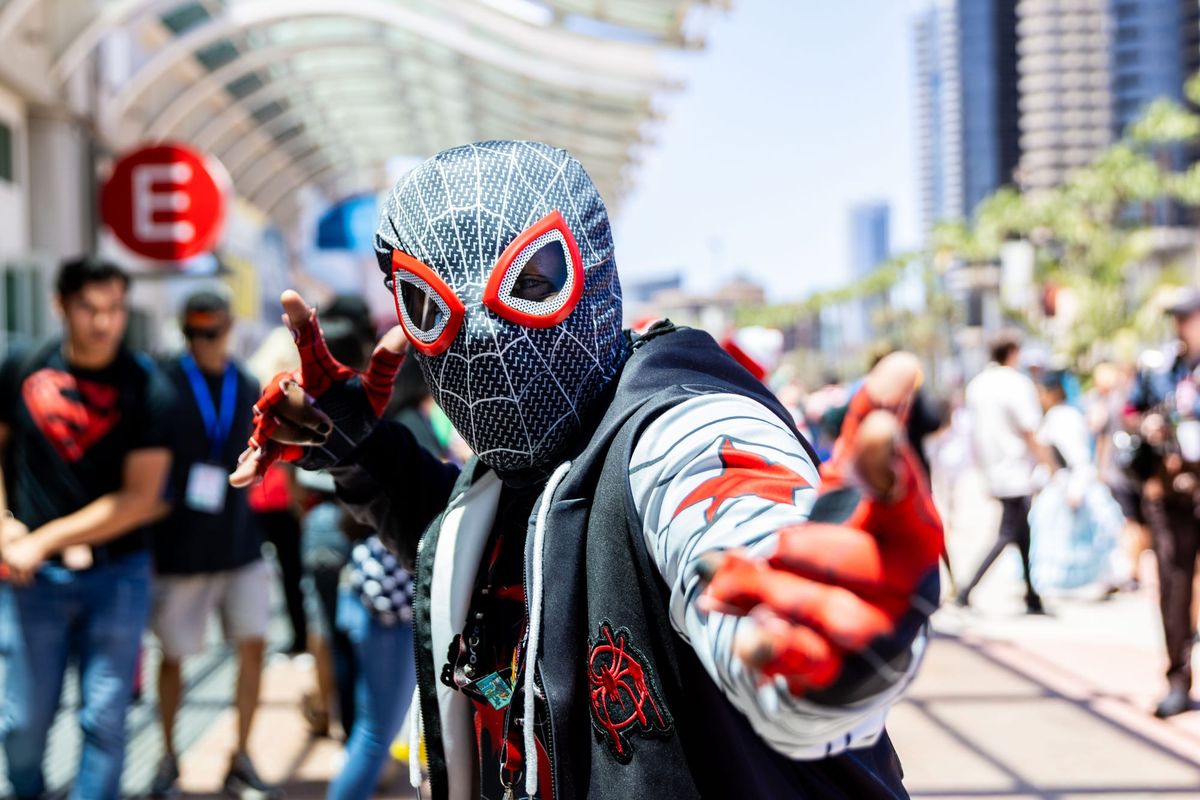 A man in a spider-man costume poses outside a convention center with people walking behind him