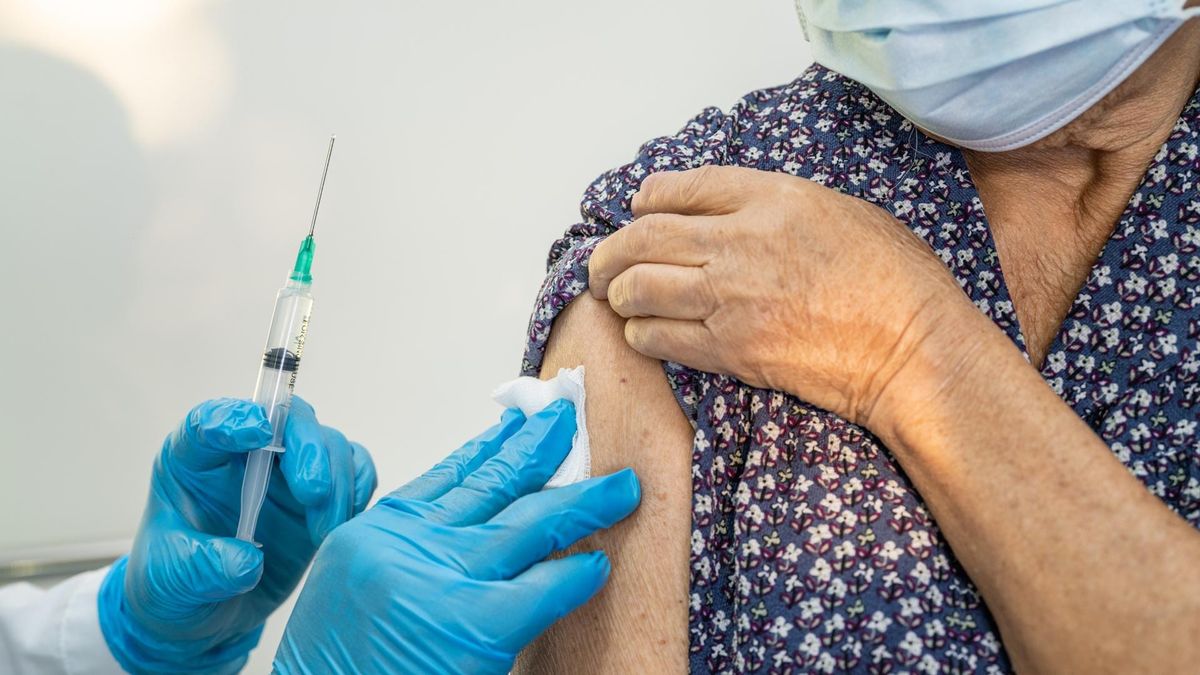 Gloved hands hold a syringe with a needle, preparing to vaccinate an arm of a masked person in a blue print shirt