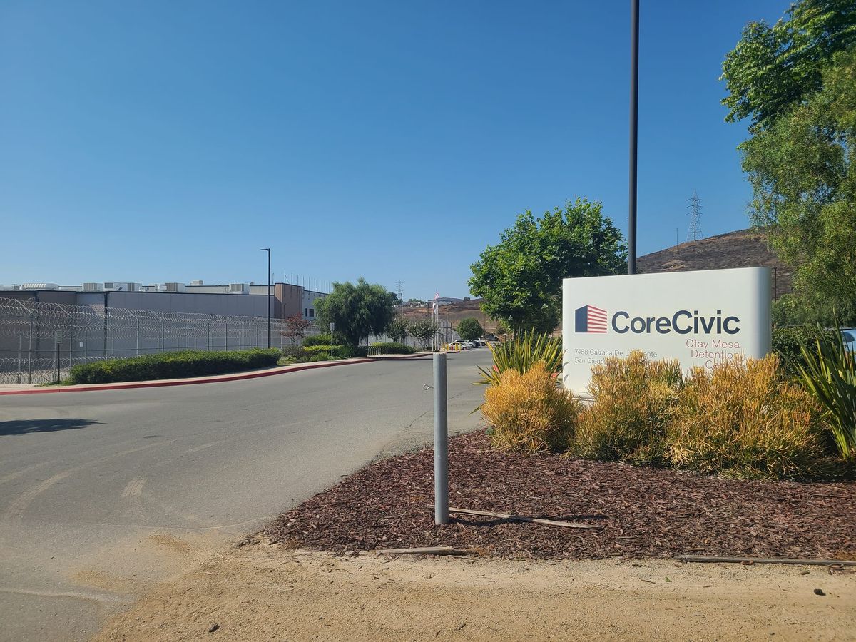 A prison-like building sits to the left of a parking lot and a sign that says CoreCivic Otay Mesa Detention Center