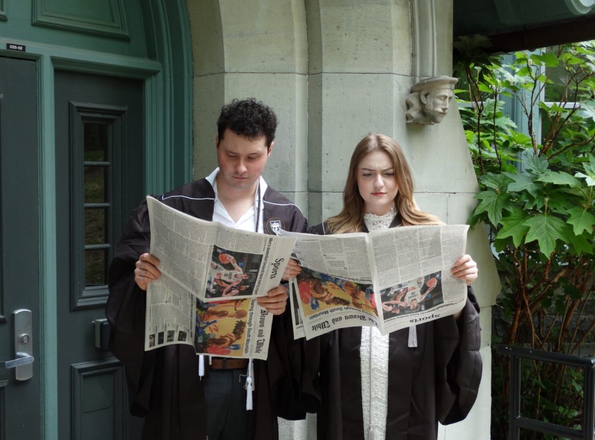 Two people in graduation robes stand outside of a college building pretending to read a newspaper, but one person has it turned upside down and the other turned to the side 