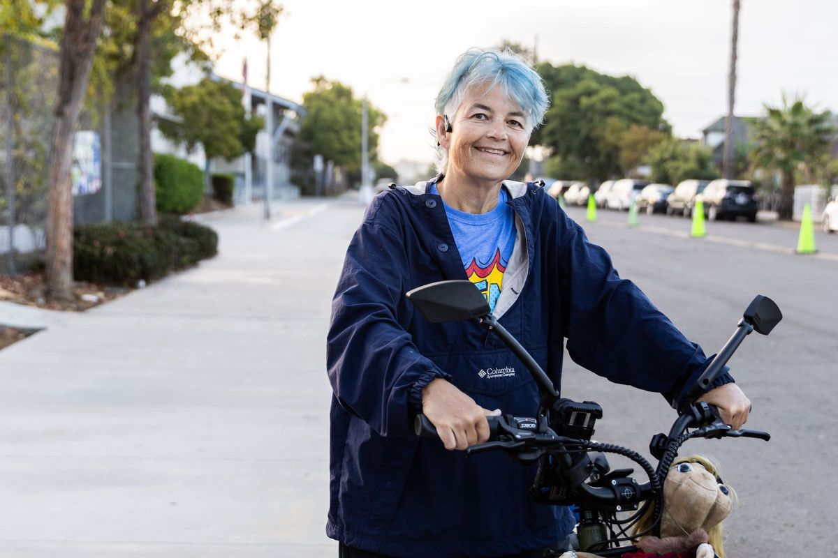 A person with blue hair and a blue jacket holds the handlebars of an e-bike with a school in the background