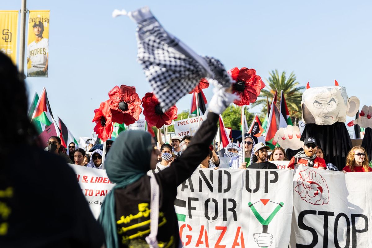 A woman waves a keffiyeh in front of a group of people marching and holding signs that read "stand up for gaza."