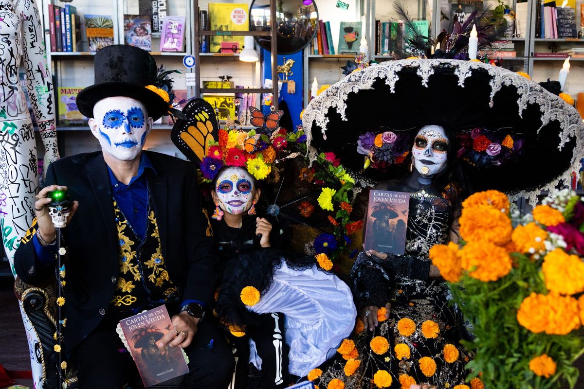A man, a little girl and a woman sit next to each other on a couch. They're wearing face paint that makes their faces look like skulls. The man is wearing a black velvet suit and a top hat. T