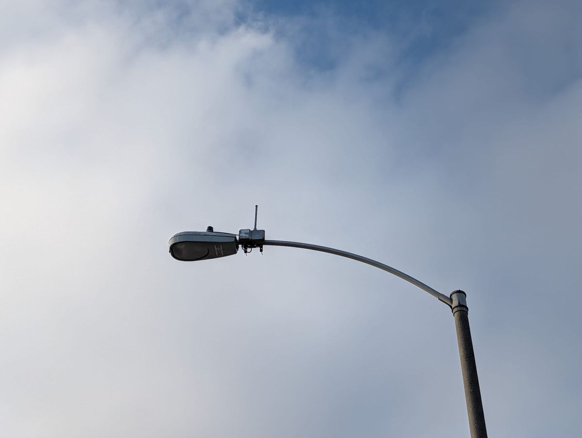 A street light with a small box with a long stick at the top looms against a cloudy sky