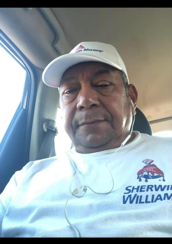 A man in a white shirt and hat sits in a car taking a selfie