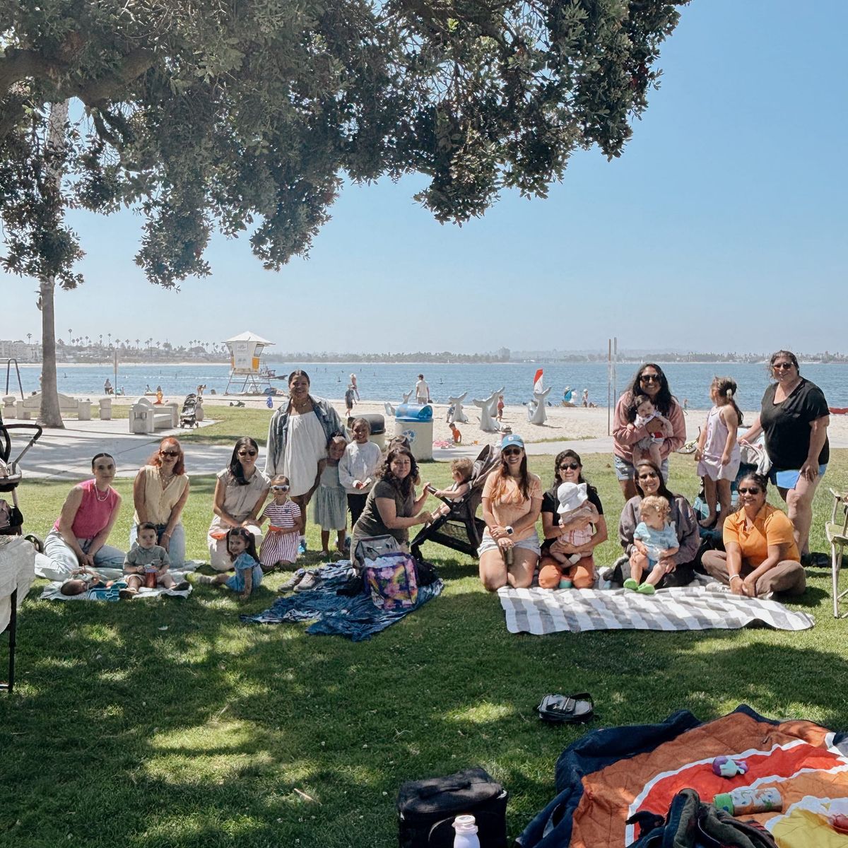 A group of moms and children sit at a park by the beach.
