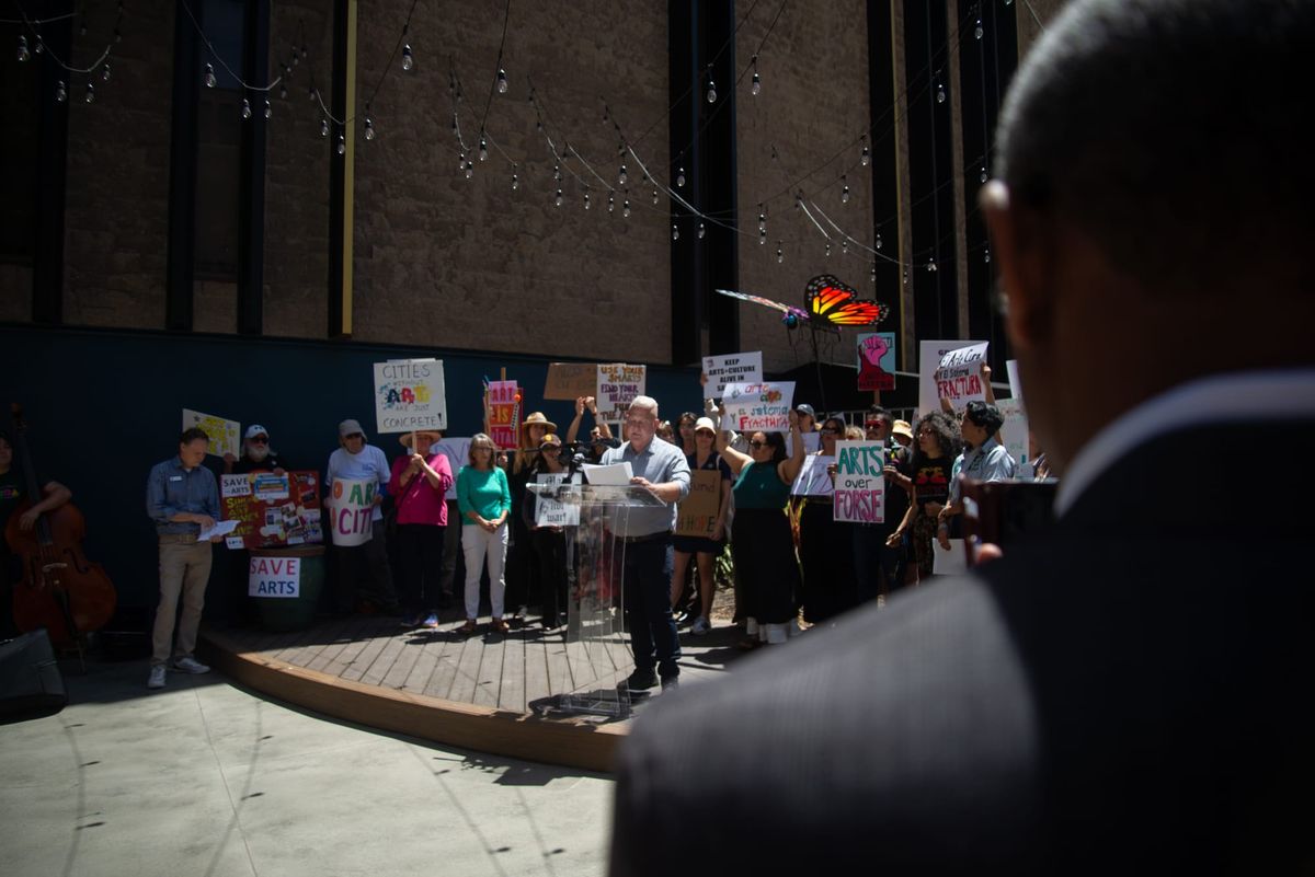 A man with blond hair stands at a podium surrounded by people with colorful signs