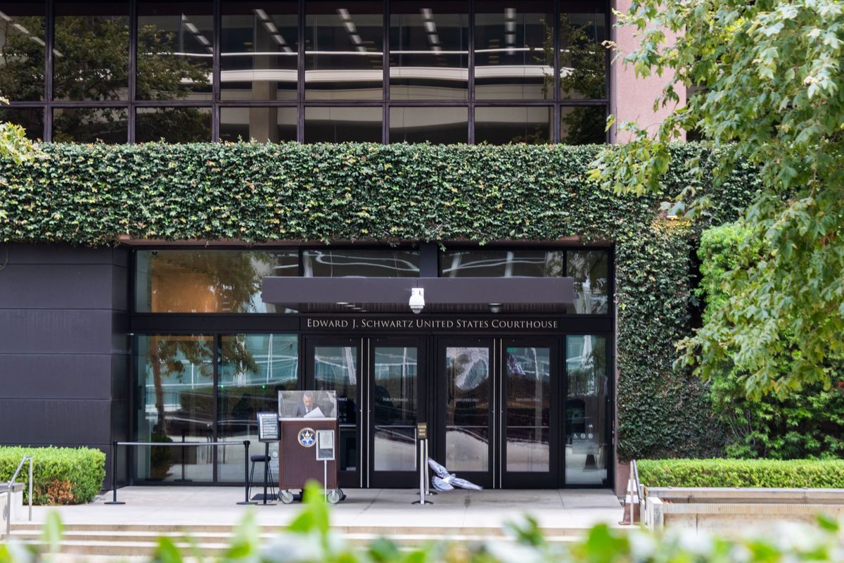 A pigeon takes off in front of a brown building with ivy growing on it and glass doors with a guard out front