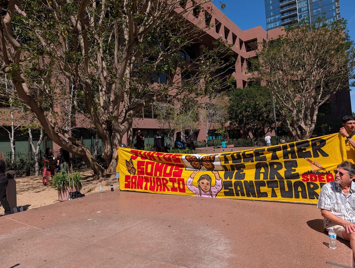 People hold a yellow sign that says juntos somos santuario in red and together we are sanctuary in black. in the background is a brick building