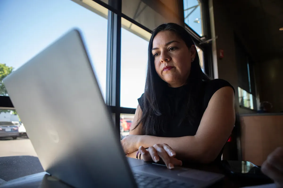 A woman in a black shirt sits in front of a laptop computer next to a large window