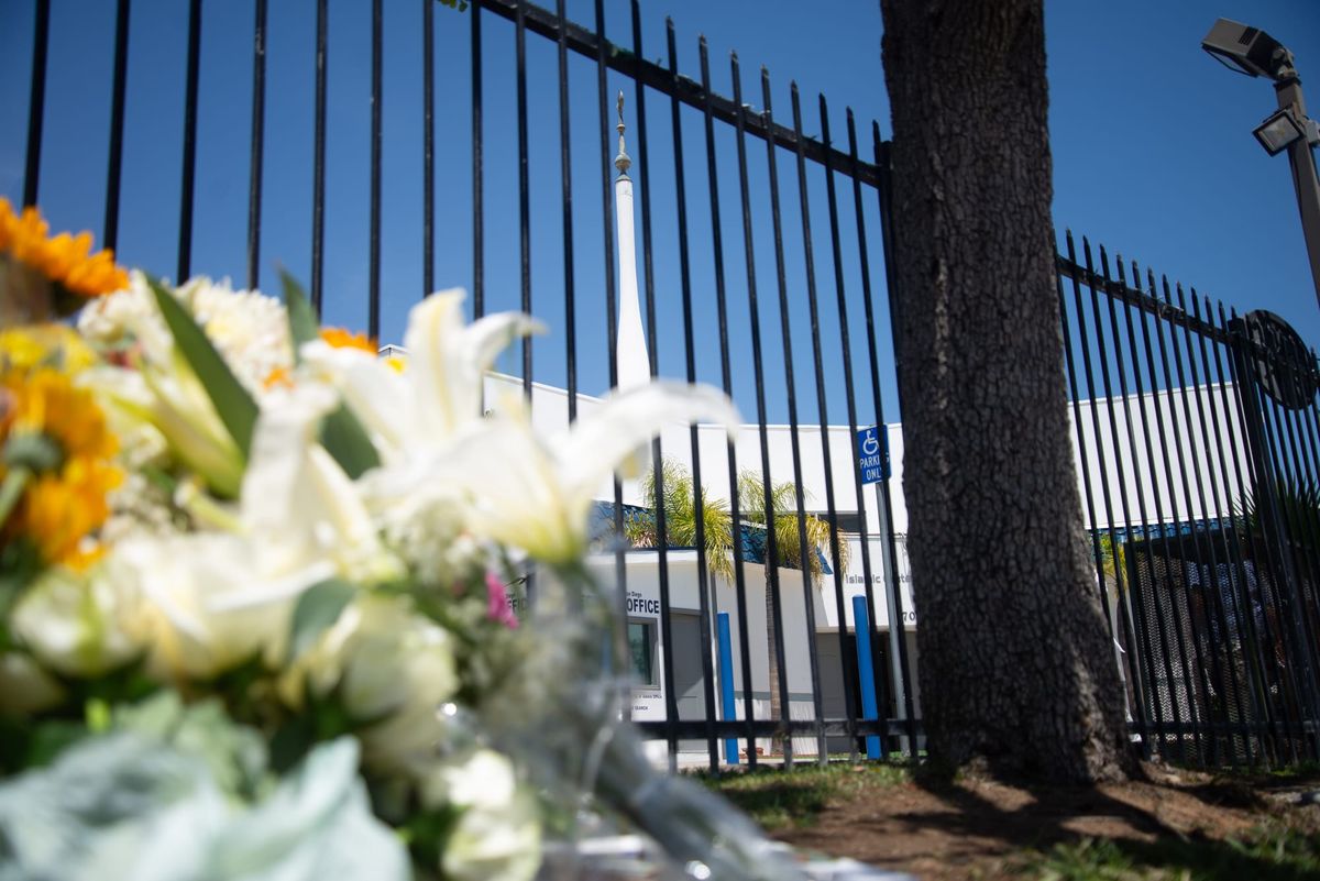 Flowers, mostly lilies, and a white box filled with baklava sit on the ground next to a palm tree in front of a mosque.