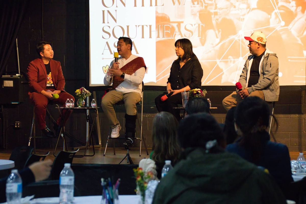 Four people sit on a stage, while one talks into a microphone. The back of people's heads can be seen watching the people on stage. 
