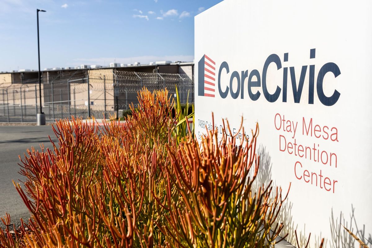 A succulent plant grows next to a sign that says CoreCivic Otay Mesa Detention Center with a prison-like building in the background