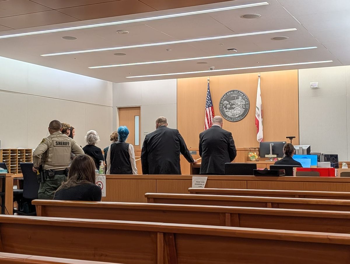 a court room with several women and two men standing facing away from the camera and a bailiff behind them with a sheriff deputy uniform on