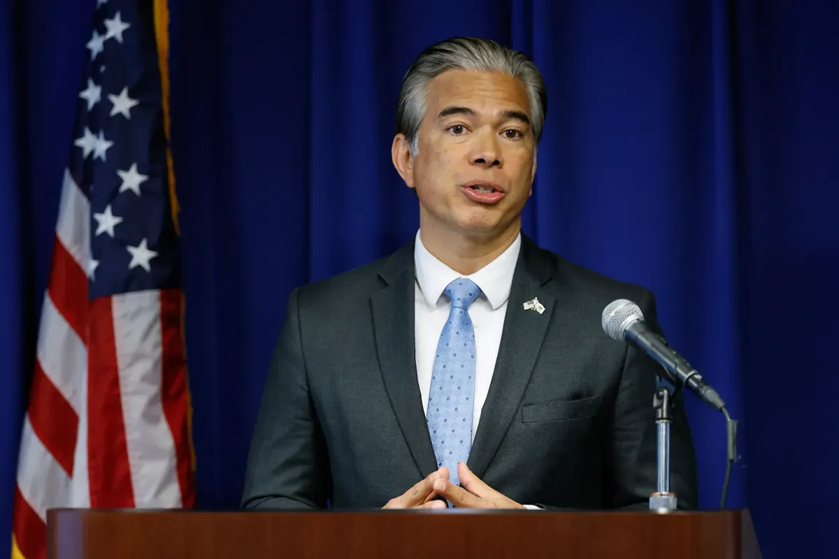 a man in a suit stands at a podium in front of a blue curtain and an american flag