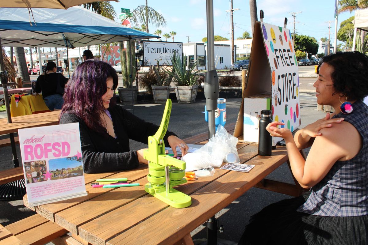 Two people are sitting at a table speaking to each other while doing arts and crafts. 