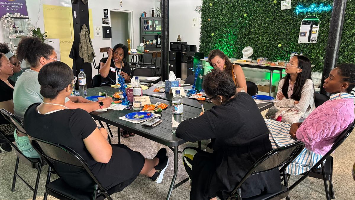 People sit around a table in an event space talking and taking notes with a green fplant wall in the background