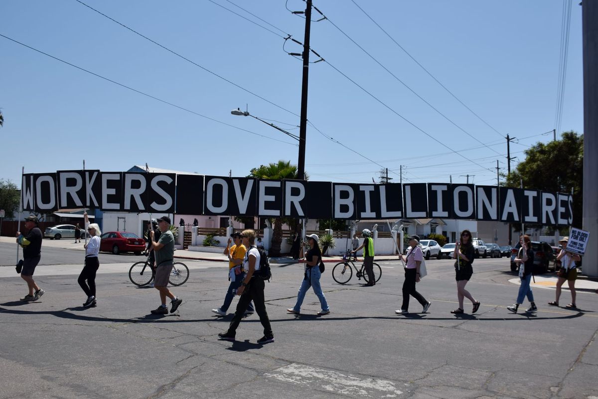 People walk in a protest holding a long sign that reads "Workers over billionaires" 