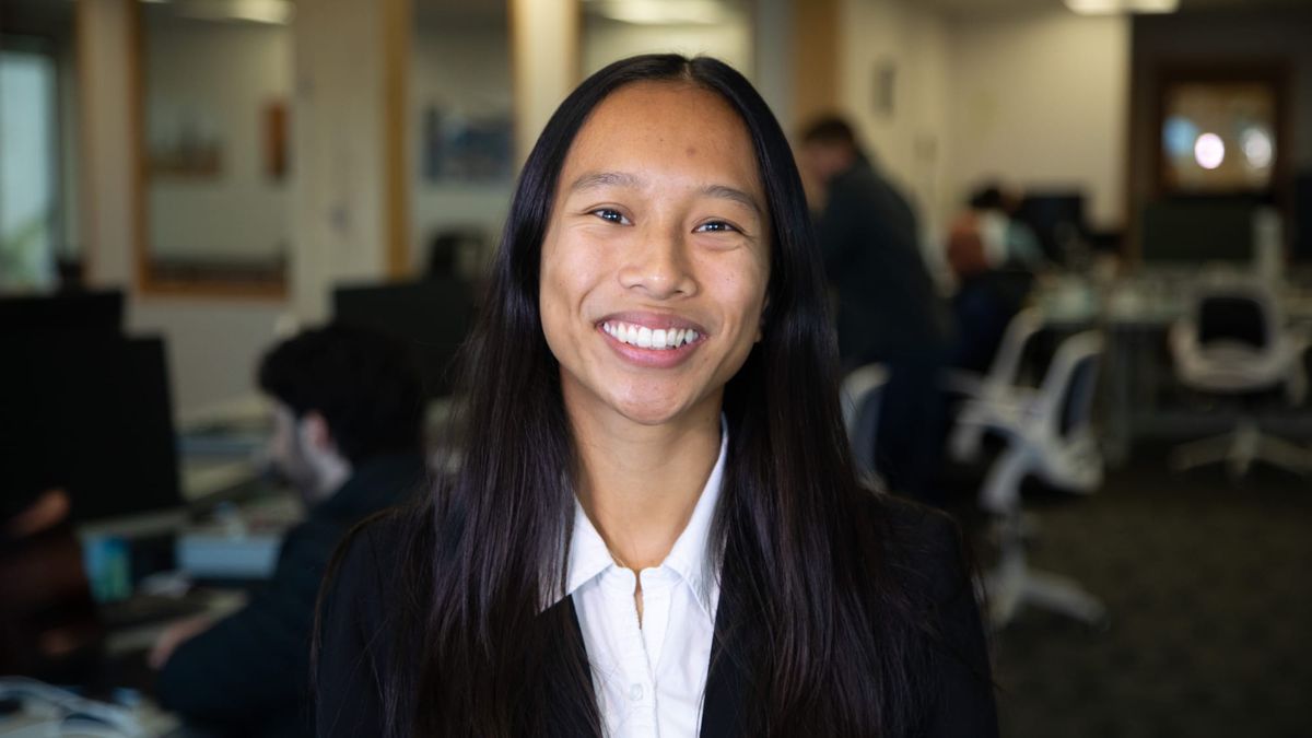 A picture of someone with straight black hair smiling in front of office workers and desks 