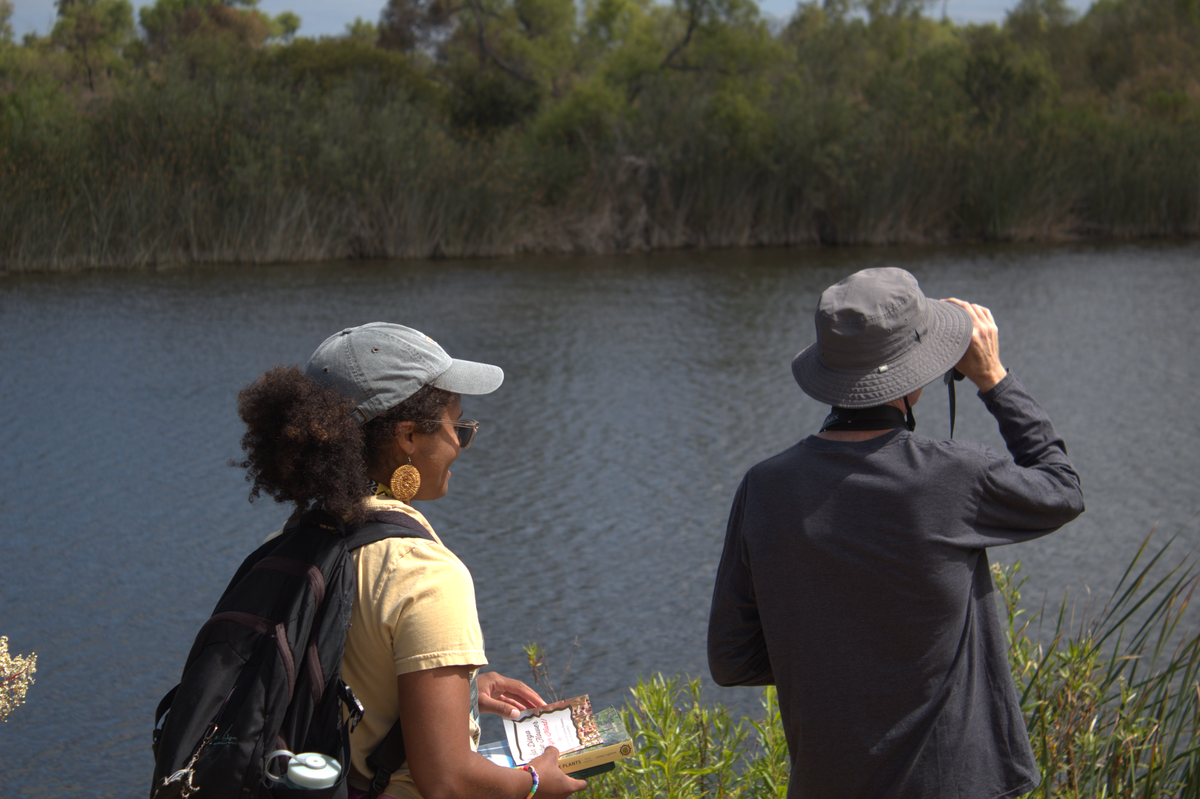 One person with a booklet in hand and another with binoculars to their eyes face a small pond with trees on the shore of the other side. 
