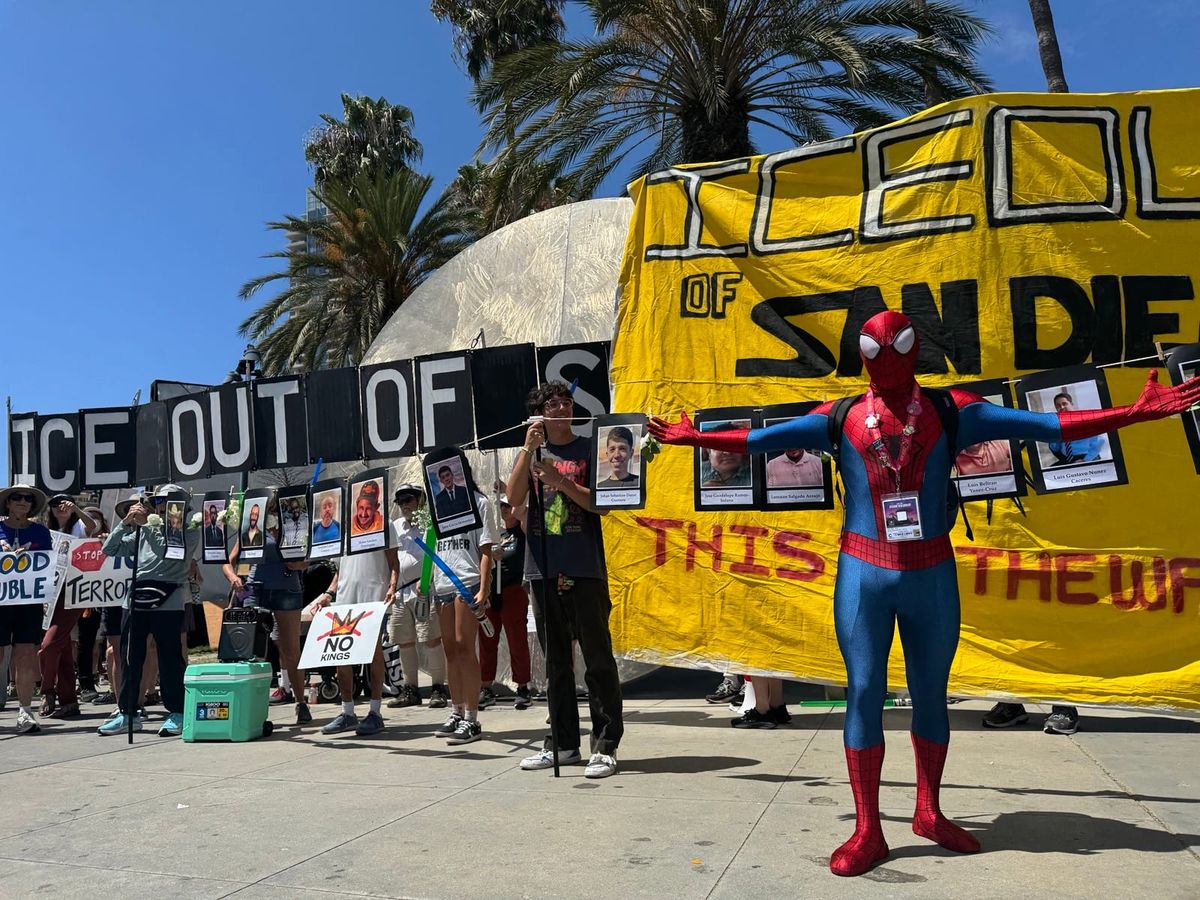 Spiderman stands in front of an ICE out of SD sign and photos of people killed by ICE